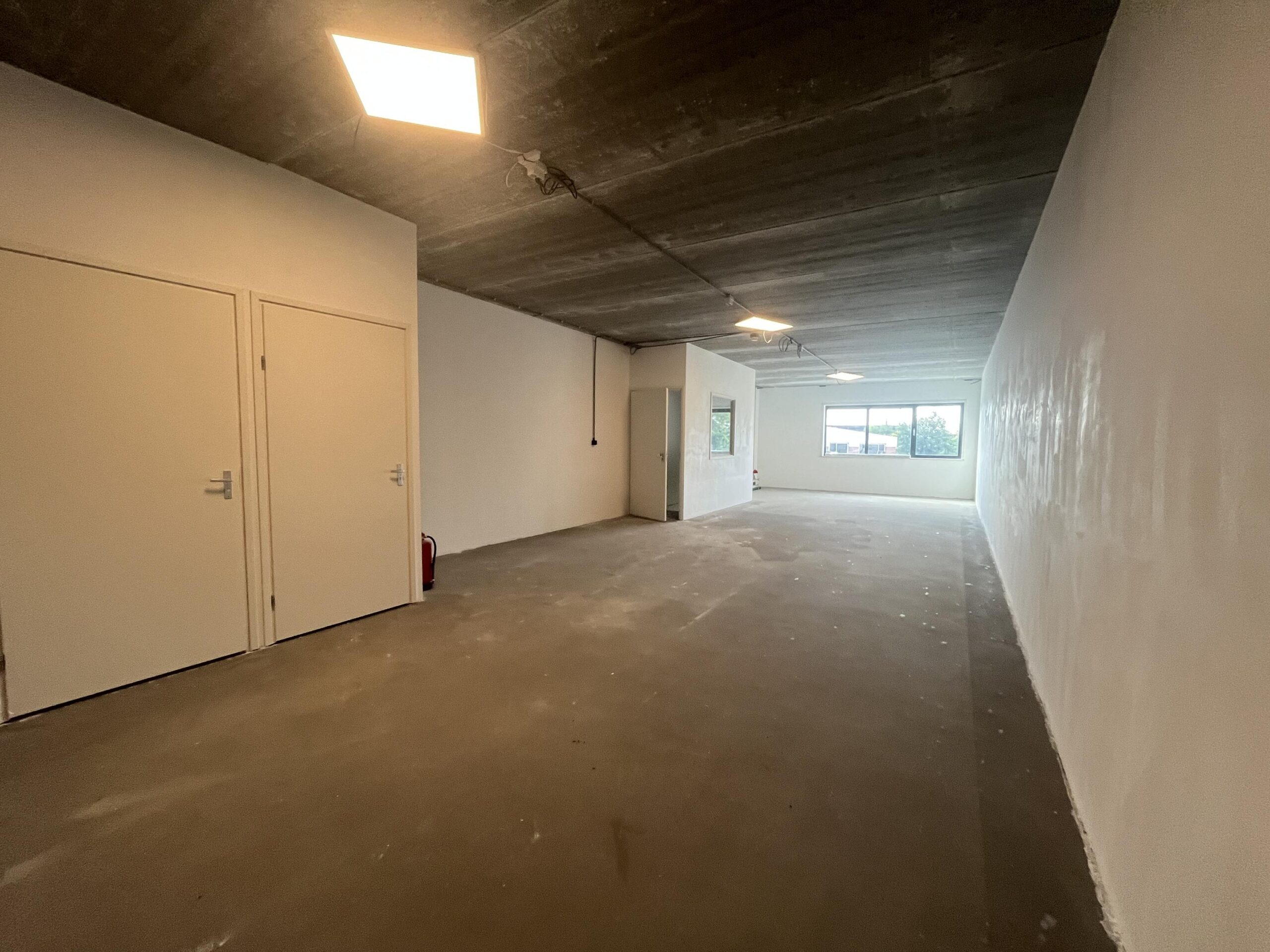 Lege, rechthoekige ruimte met witte muren, een betonnen vloer en plafond, enkele deuren en een raam aan het einde dat uitzicht biedt naar buiten.