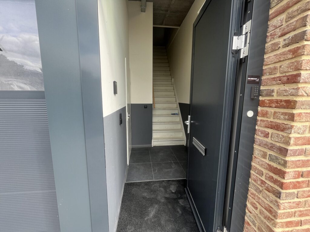 Een openstaande voordeur met een zicht op een smalle hal en een trap naar boven.