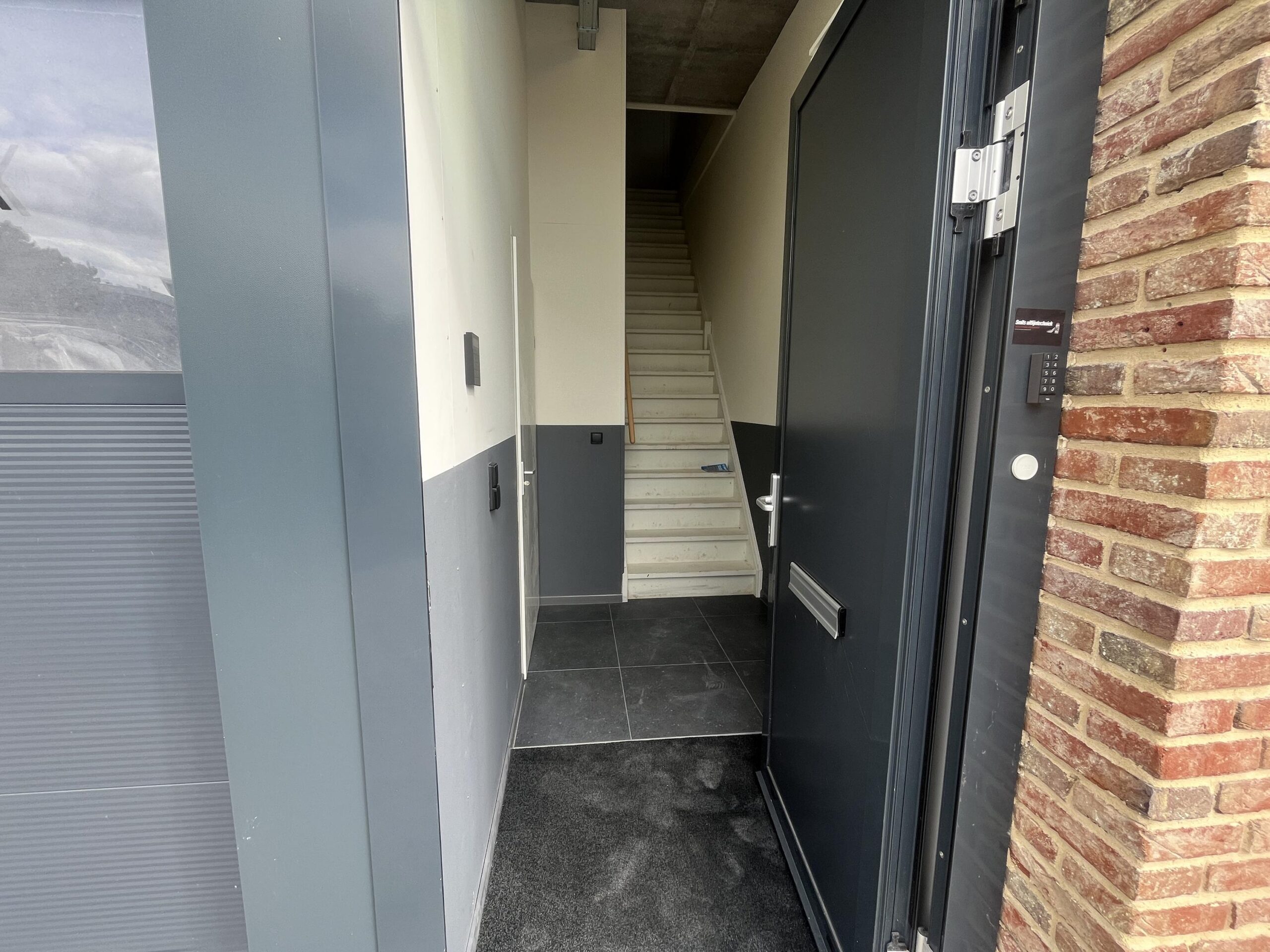 Een openstaande voordeur met een zicht op een smalle hal en een trap naar boven.