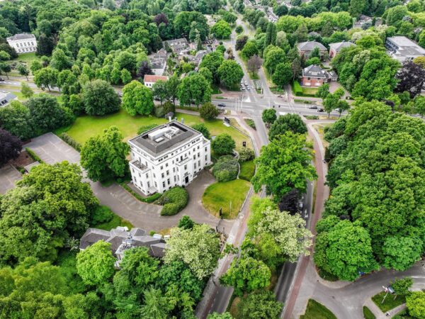 Luchtfoto van een wit stadsvilla aan de Utrechtseweg in een groene, boomrijke omgeving met omliggende wegen en woningen.