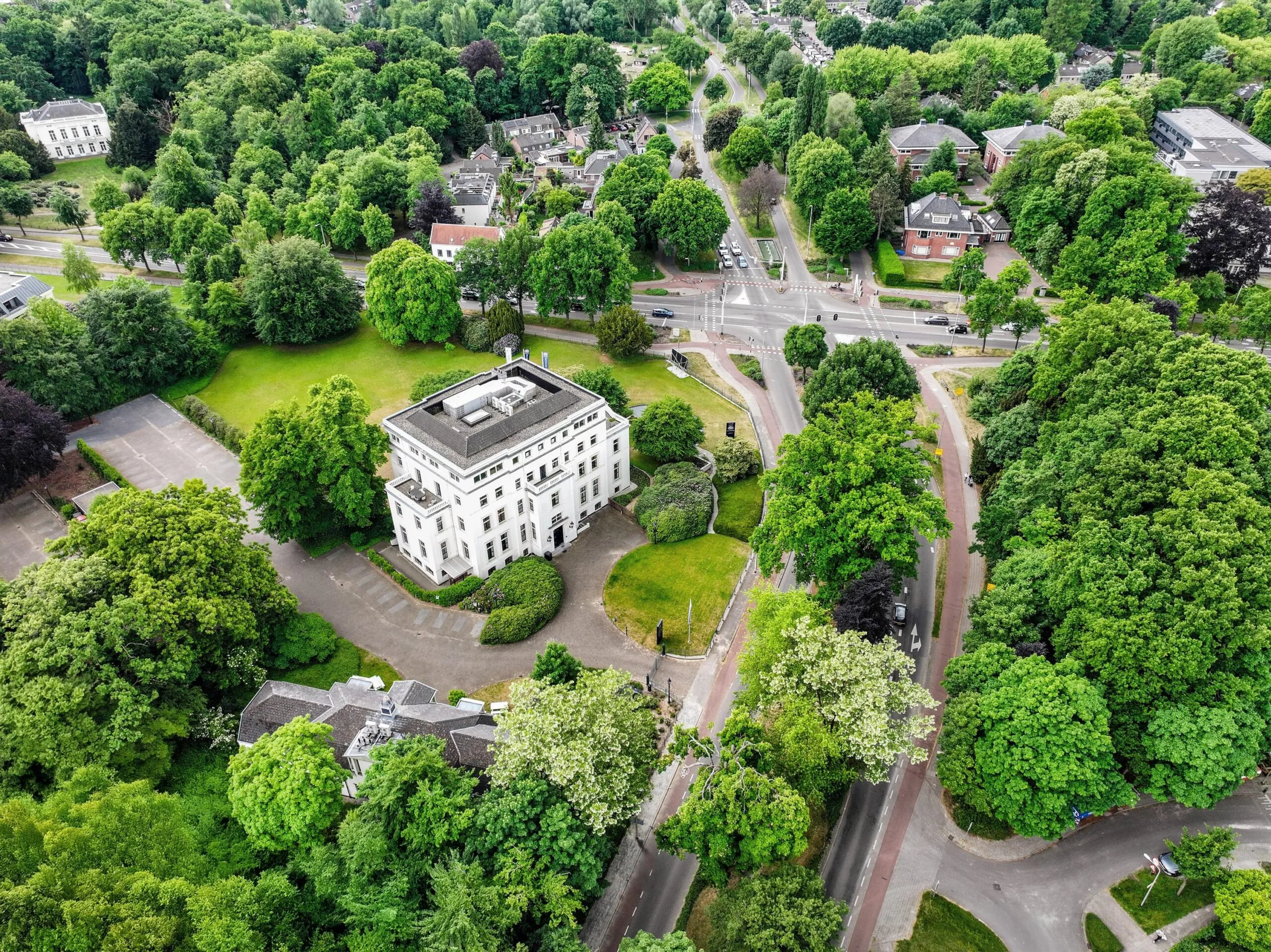 Luchtfoto van een wit stadsvilla aan de Utrechtseweg in een groene, boomrijke omgeving met omliggende wegen en woningen.