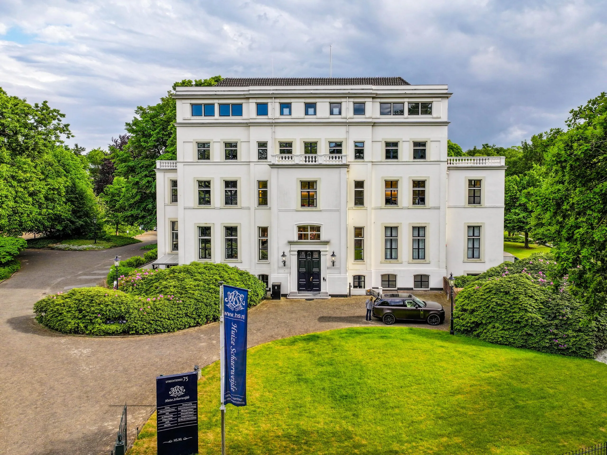 Wit herenhuis Huis Schuylenburg aan de Utrechtseweg, omgeven door groen en een oprijlaan met auto.