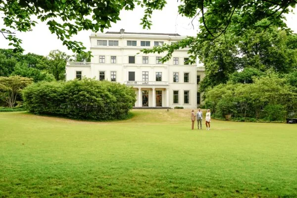 Drie mensen staan op een groot gazon voor een wit, klassiek herenhuis aan de Utrechtseweg, omgeven door groen.