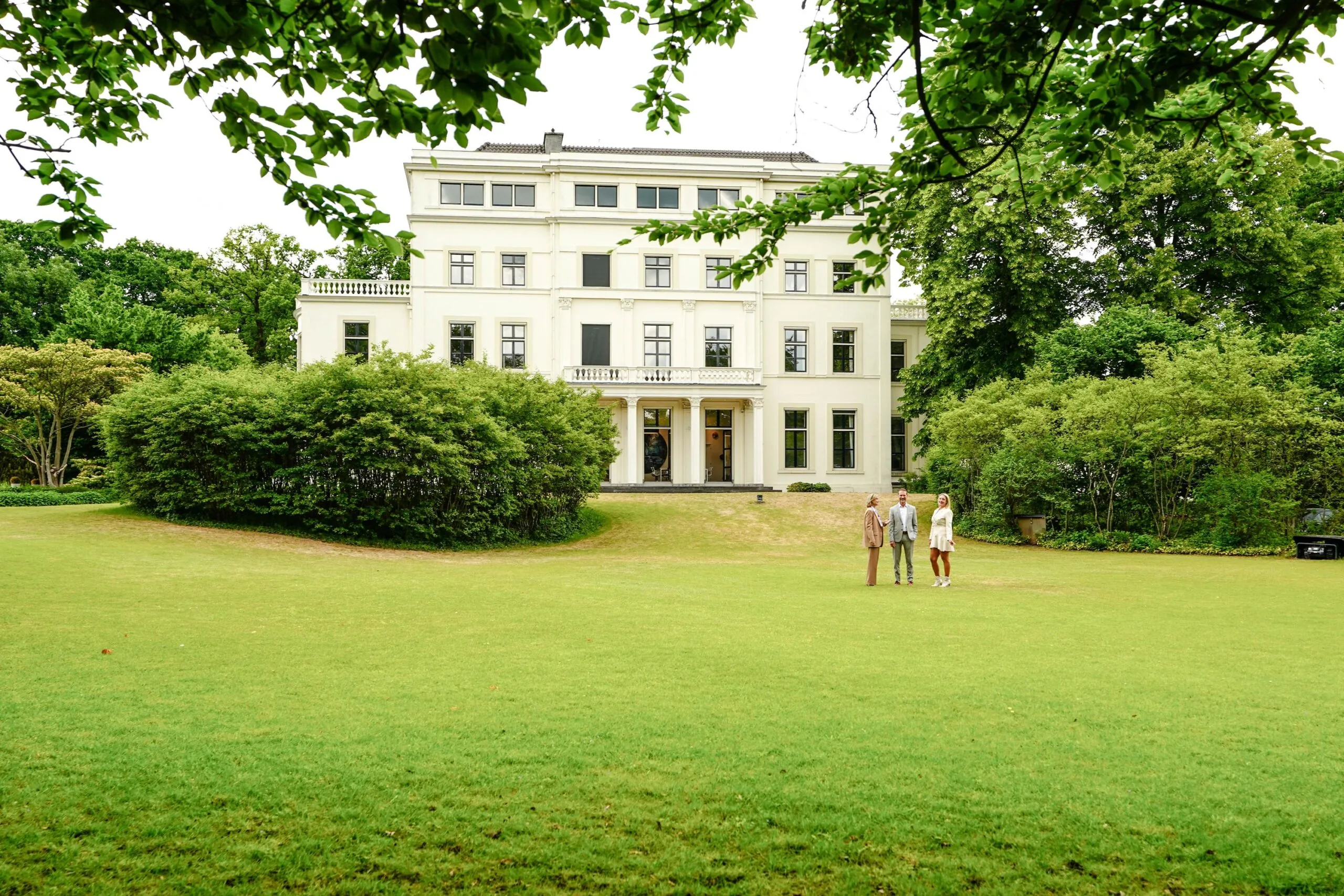 Drie mensen staan op een groot gazon voor een wit, klassiek herenhuis aan de Utrechtseweg, omgeven door groen.