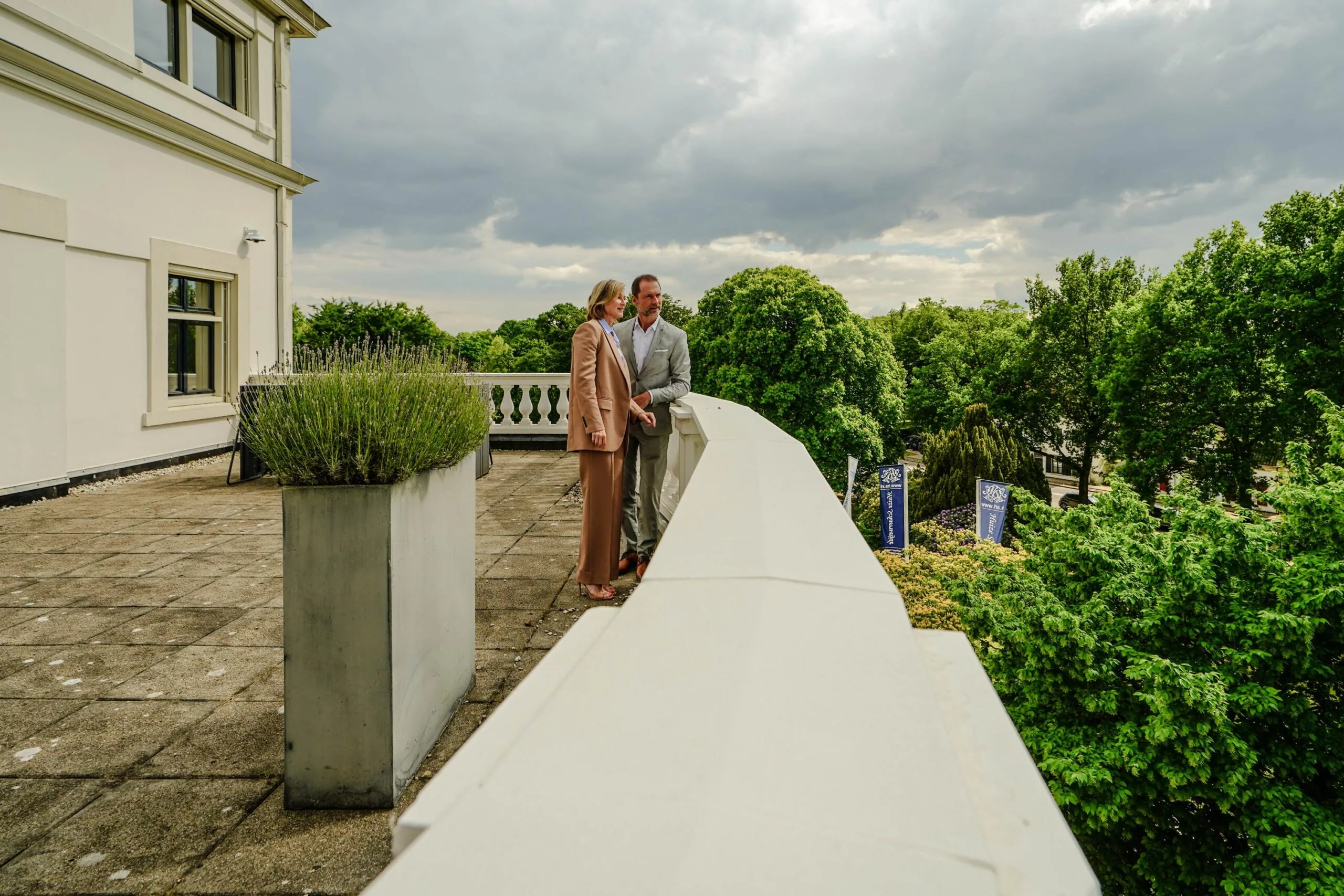 Twee mensen staan op een balkon van een wit gebouw aan de Utrechtseweg, uitkijkend over een groene, boomrijke omgeving.