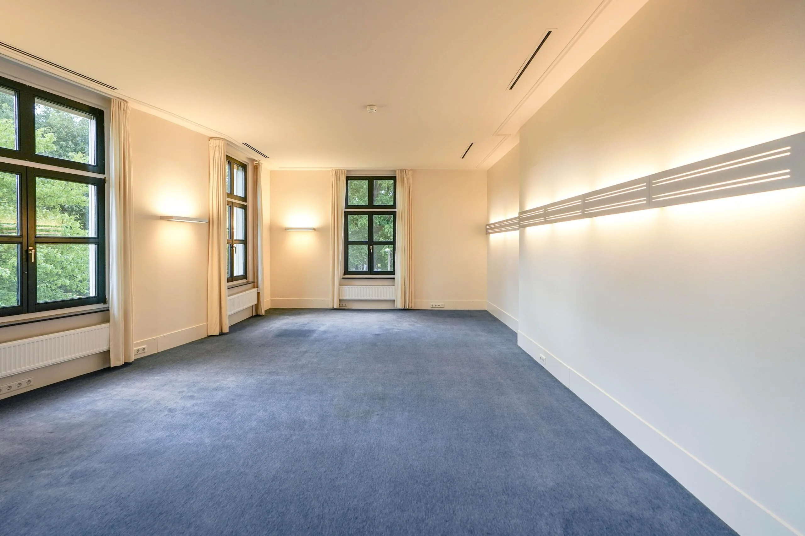 Lichte, ruime kamer aan de Utrechtseweg met blauw tapijt, grote ramen en moderne wandverlichting.
