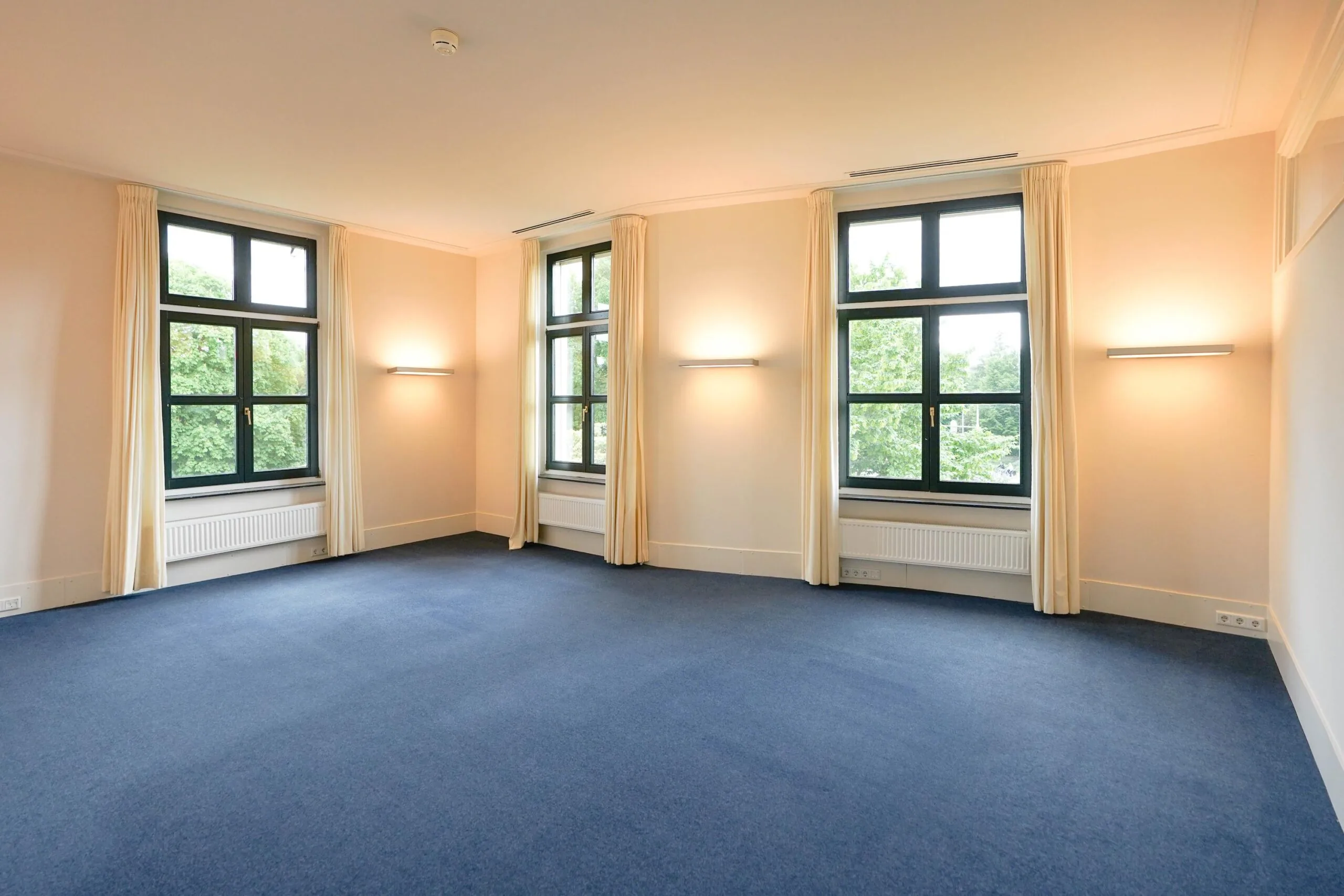 Lichte lege kamer aan de Utrechtseweg met drie grote ramen, blauwe vloerbedekking en crèmekleurige gordijnen.