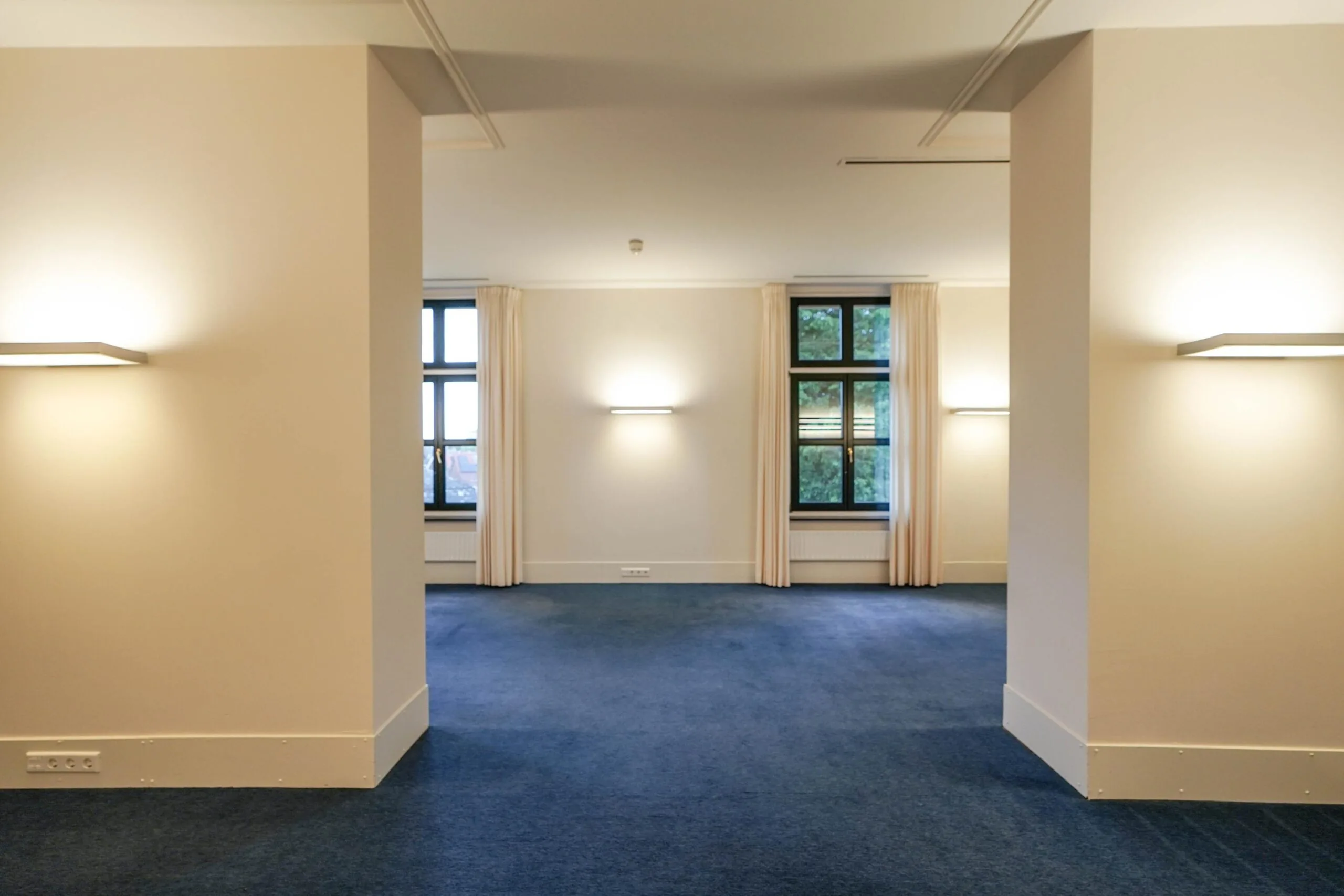 Lege kantoorruimte aan de Utrechtseweg met blauwe vloerbedekking, beige muren en grote ramen met lichte gordijnen.