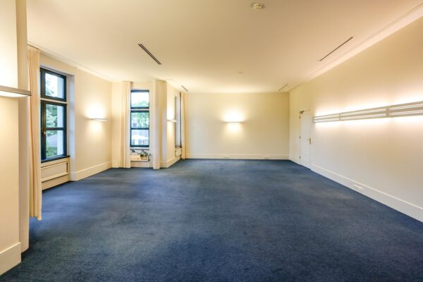 Ruime, lichte kamer aan de Utrechtseweg met grote ramen, blauwe vloerbedekking en moderne wandverlichting.