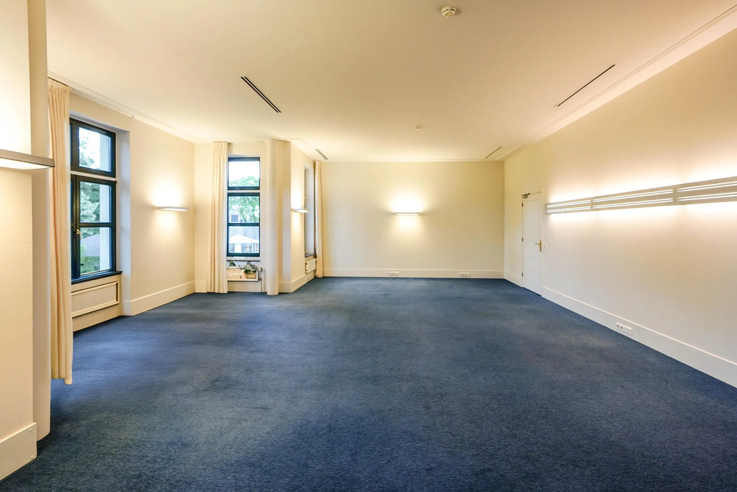 Ruime, lichte kamer aan de Utrechtseweg met grote ramen, blauwe vloerbedekking en moderne wandverlichting.