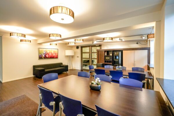 Moderne vergaderruimte aan de Utrechtseweg met houten tafel, blauwe stoelen, kunst aan de muur en ingebouwde keukenfaciliteiten.