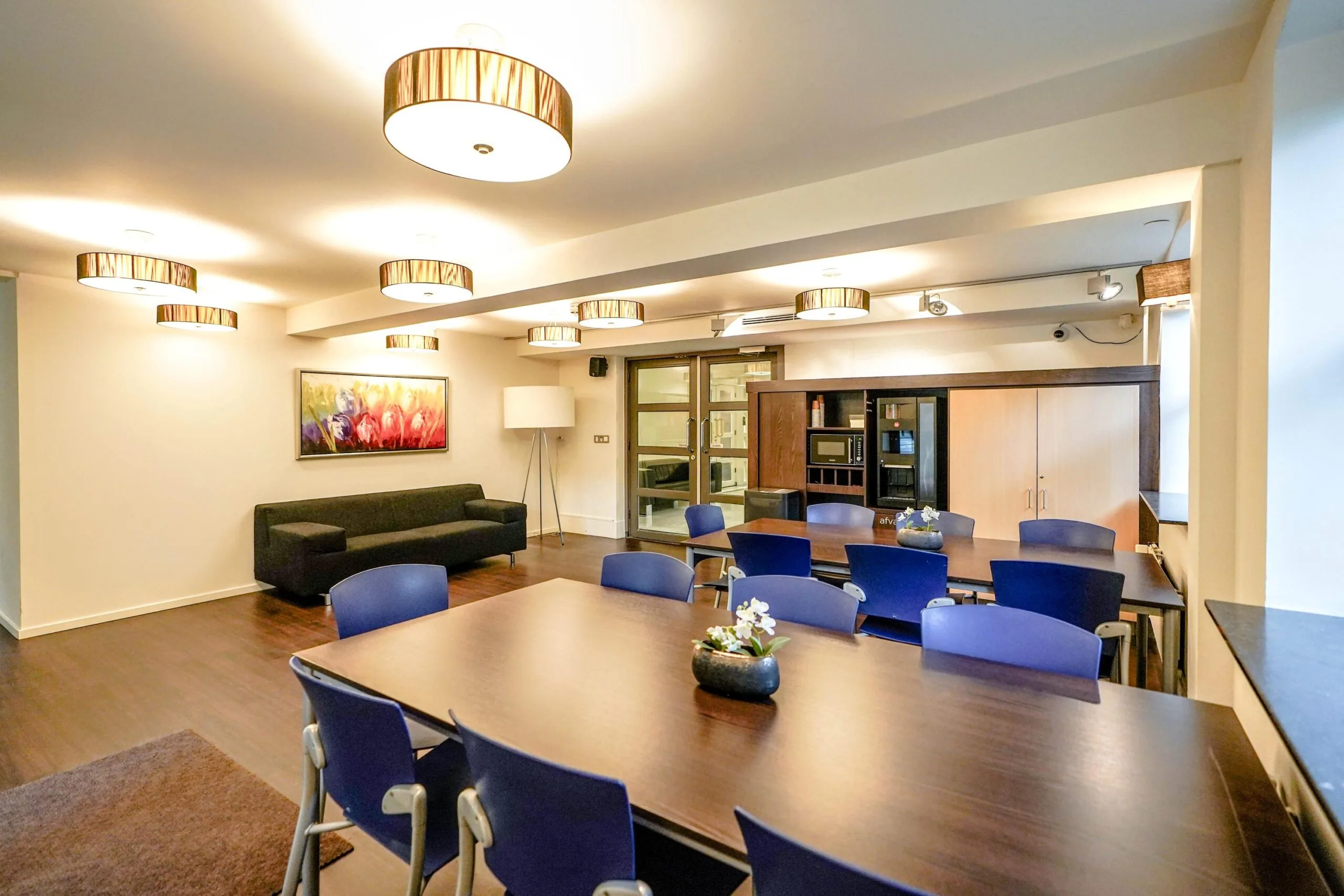 Moderne vergaderruimte aan de Utrechtseweg met houten tafel, blauwe stoelen, kunst aan de muur en ingebouwde keukenfaciliteiten.