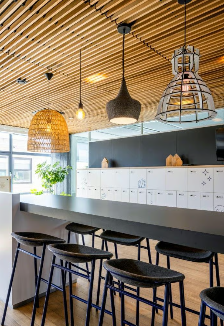 Moderne kantoorkeuken aan de Boeing Avenue met een hoge tafel, barkrukken, sfeervolle hanglampen en witte lockers op de achtergrond.