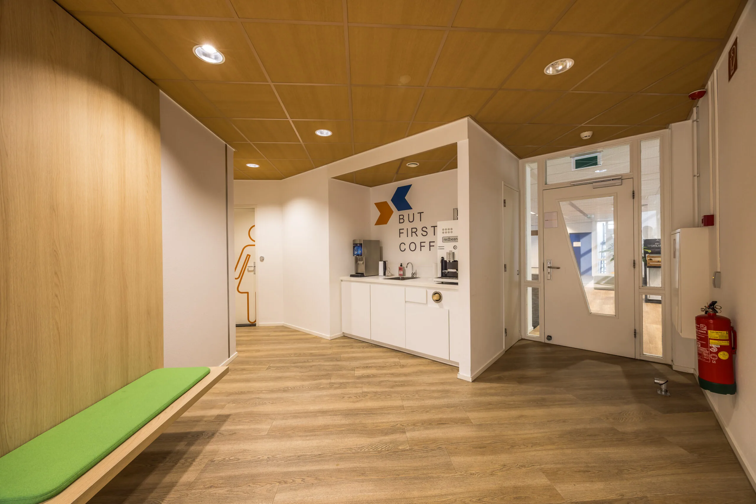 Moderne kantoorruimte met koffiehoek, groene zitbank en een toiletdeur in de gang.