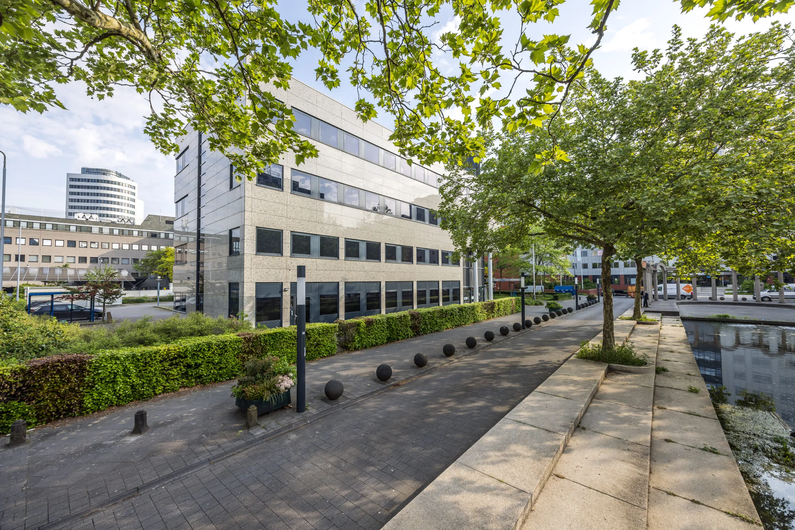 Modern kantoorgebouw aan de Varrolaan met bestrate wandelpaden, bomen en een vijver op de voorgrond.