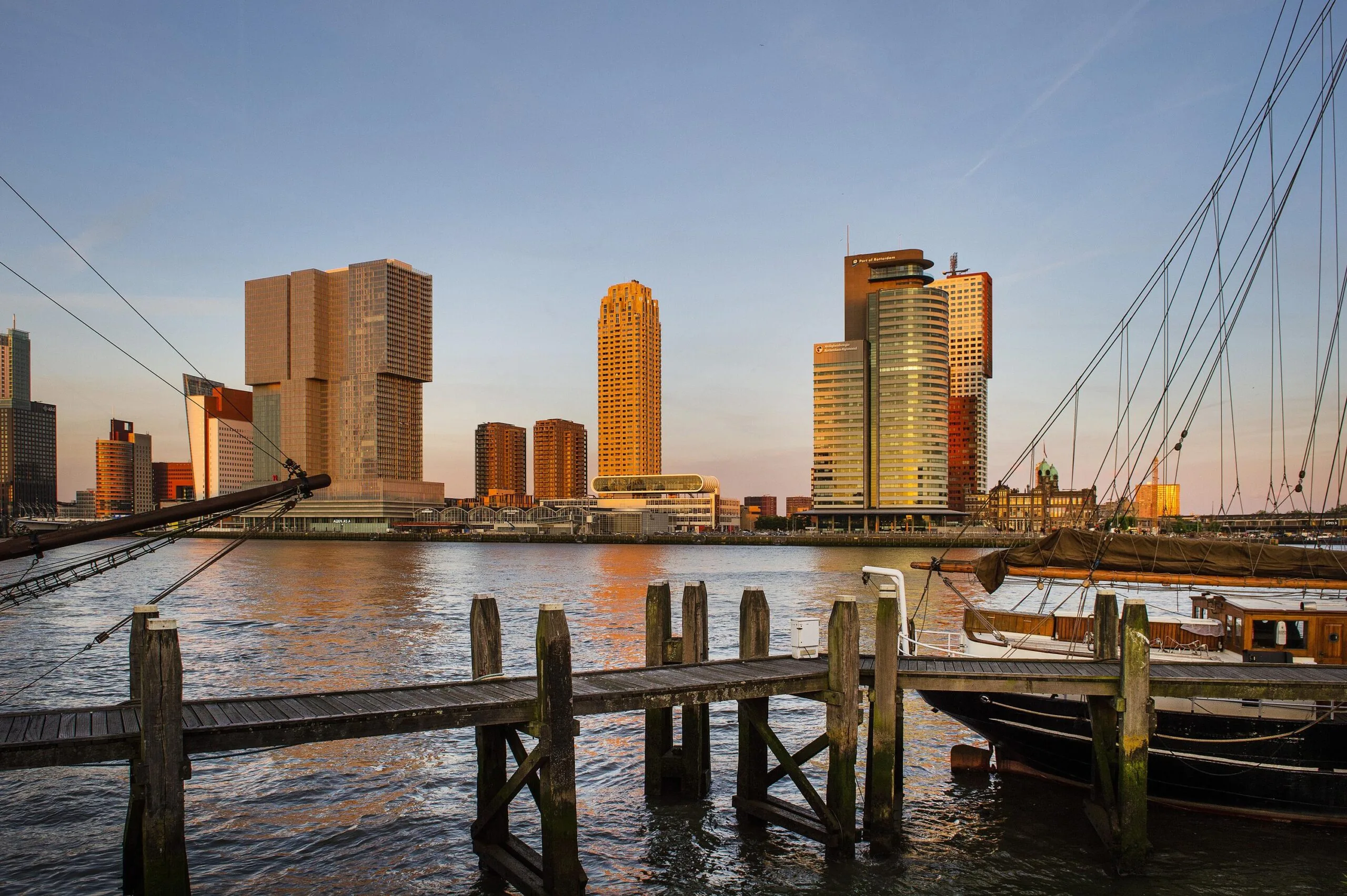 Zicht op de Wilhelminakade in Rotterdam met moderne hoogbouw en een aangemeerd zeilschip bij zonsondergang.