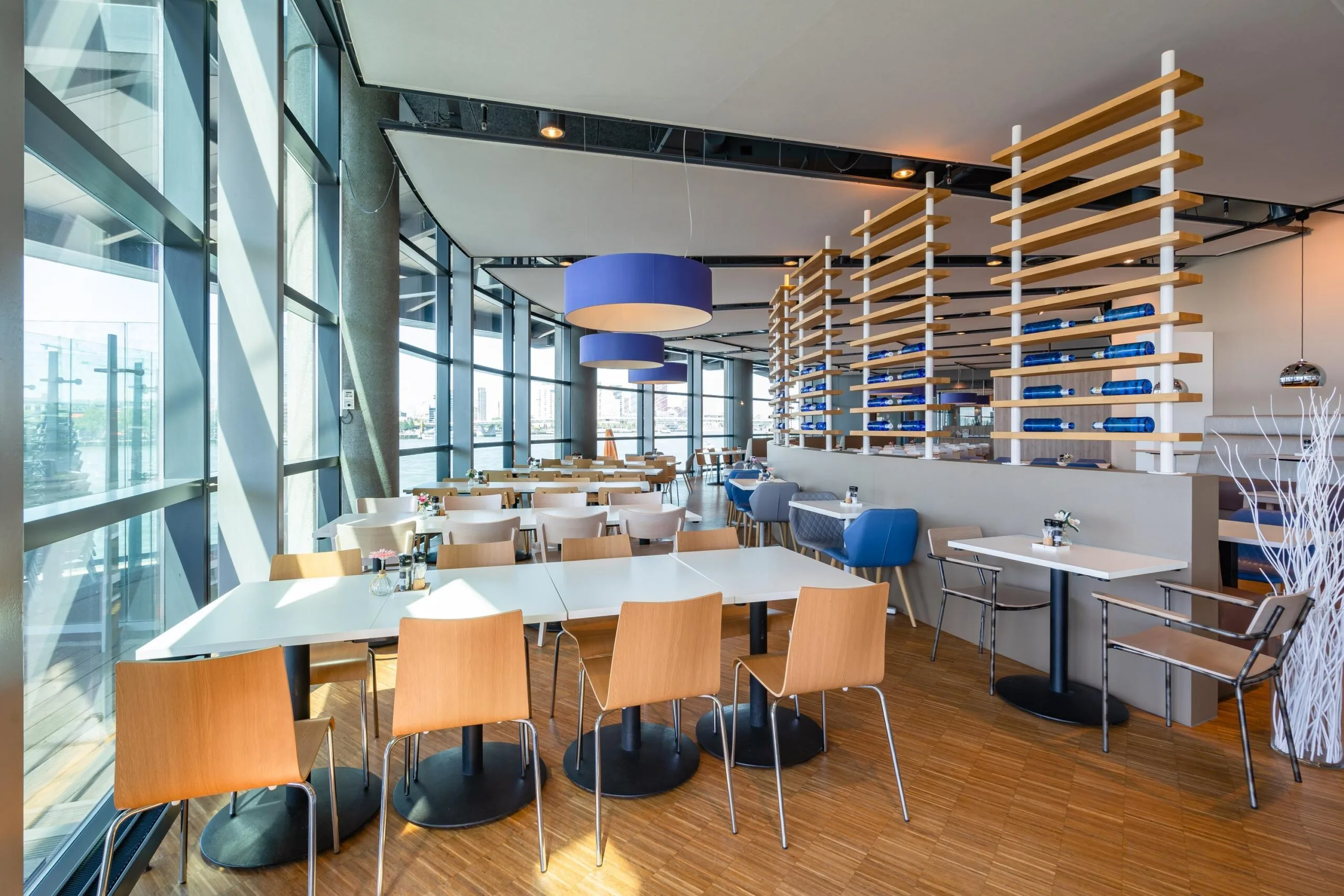 Modern restaurantinterieur aan de Wilhelminakade met grote ramen, houten stoelen en blauwe accenten.