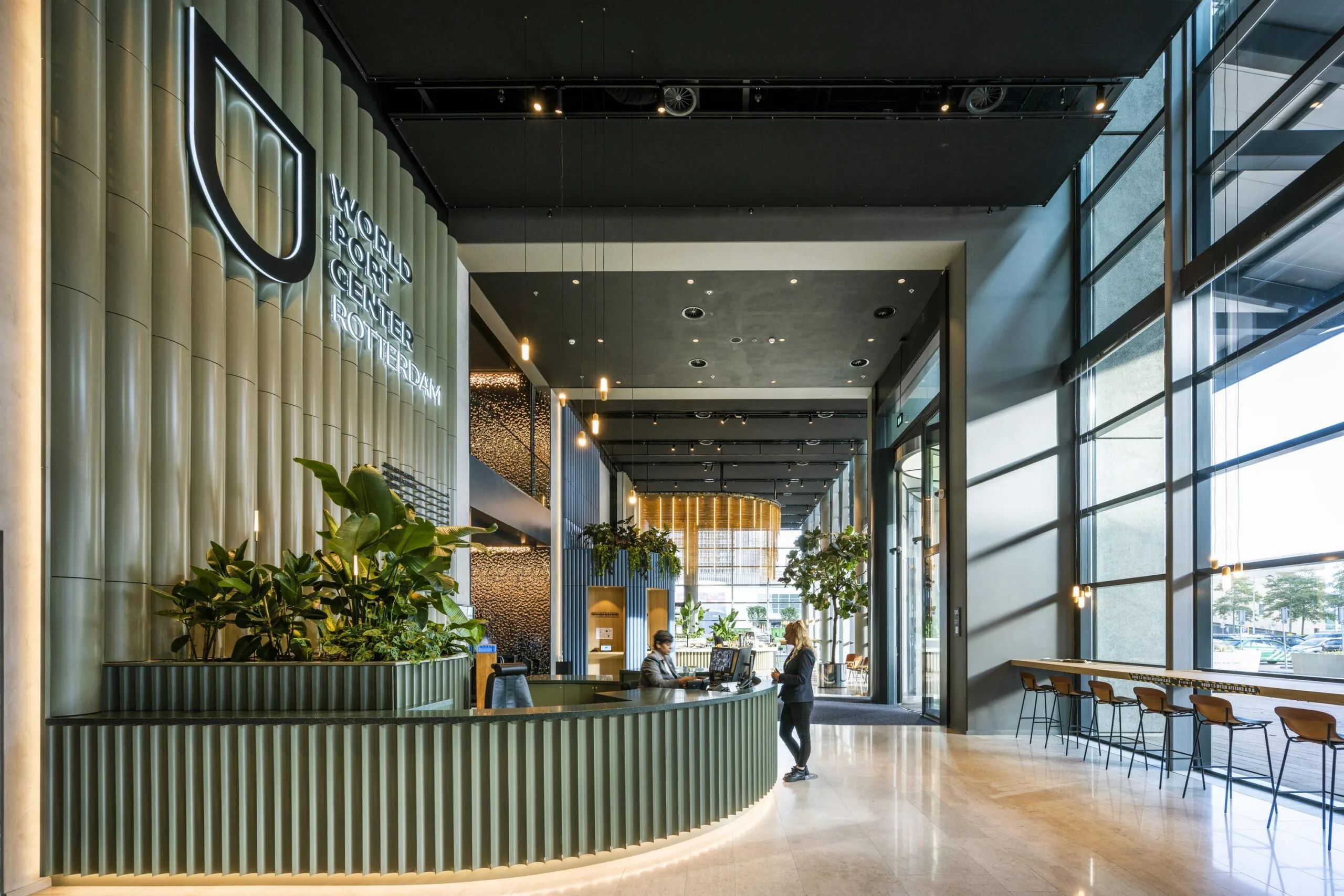 Moderne ontvangstruimte van het World Port Center Rotterdam met een groene balie, veel daglicht en planten.