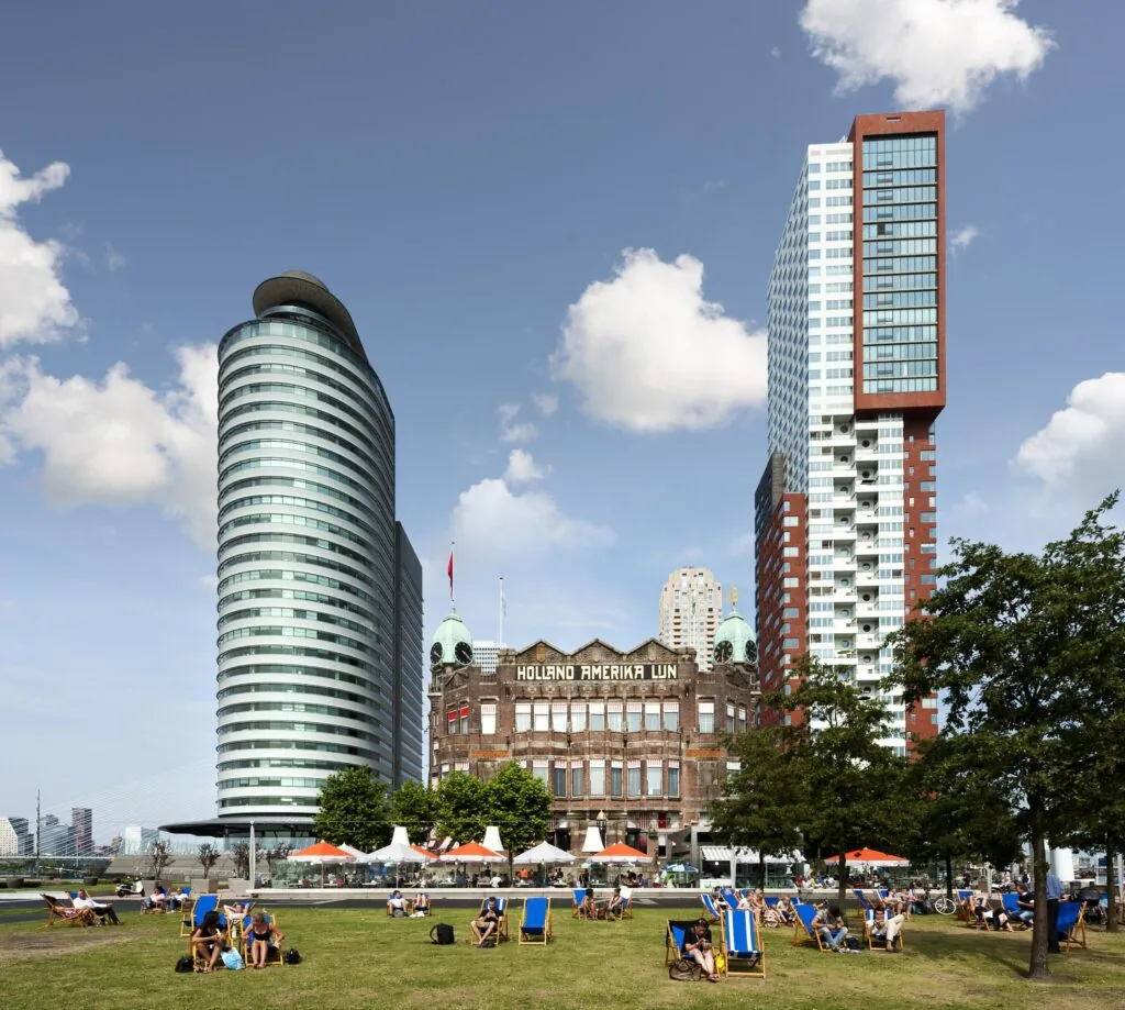 Mensen relaxen op ligstoelen in het gras aan de Wilhelminakade in Rotterdam, met op de achtergrond het gebouw van Hotel New York en moderne hoogbouw.