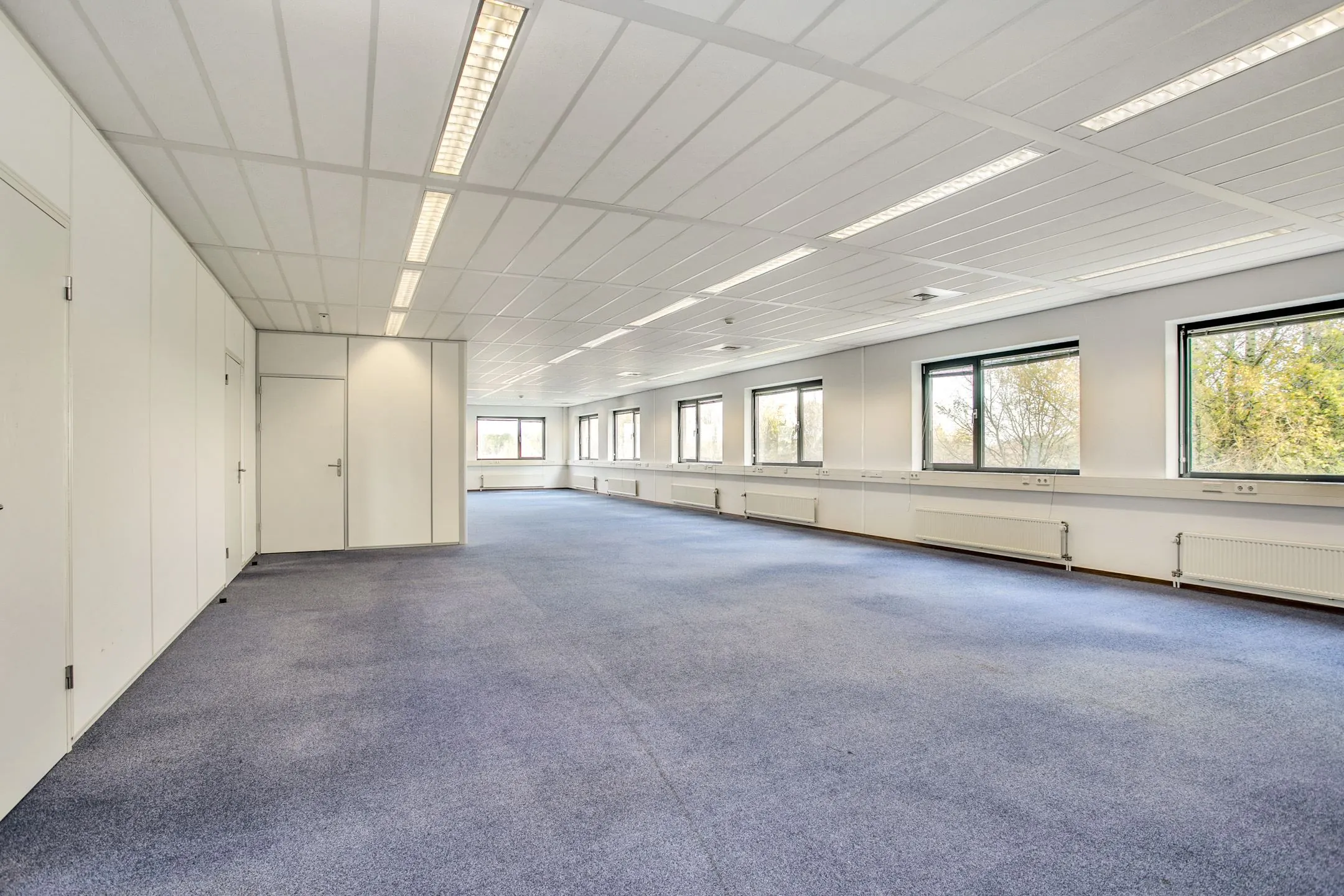 Lichte, lege kantoorruimte aan de Laan van S-Gravenmade met blauwe vloerbedekking en grote ramen.