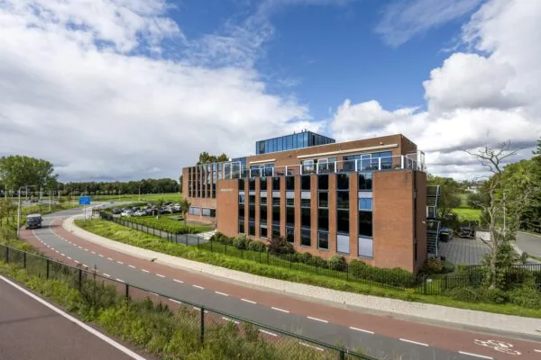 Kantoorgebouw aan de Oranjebaan met bakstenen gevel en grote ramen, gelegen naast een bochtige weg en parkeerterrein.