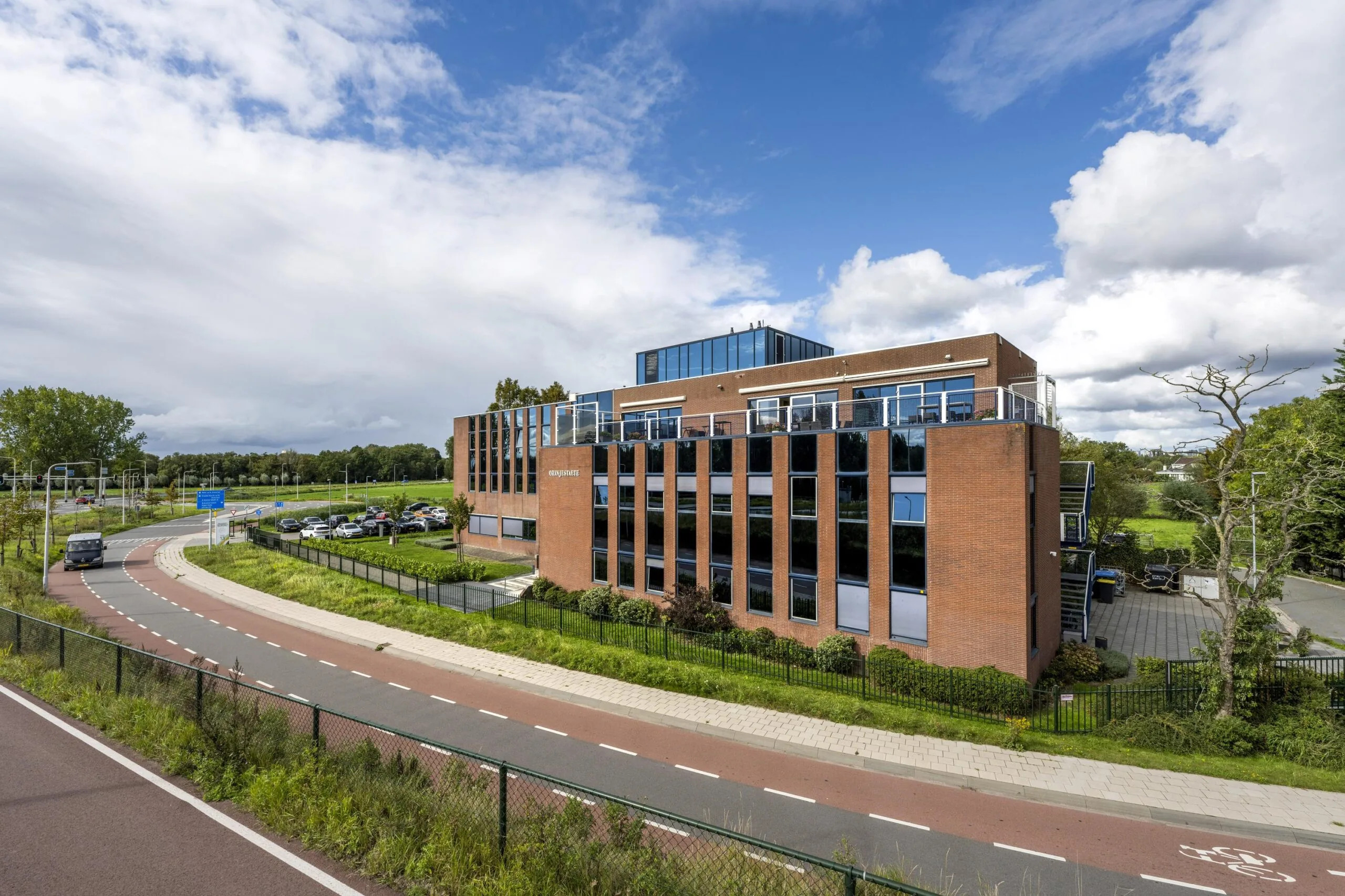 Kantoorgebouw aan de Oranjebaan met bakstenen gevel en grote ramen, gelegen naast een bochtige weg en parkeerterrein.
