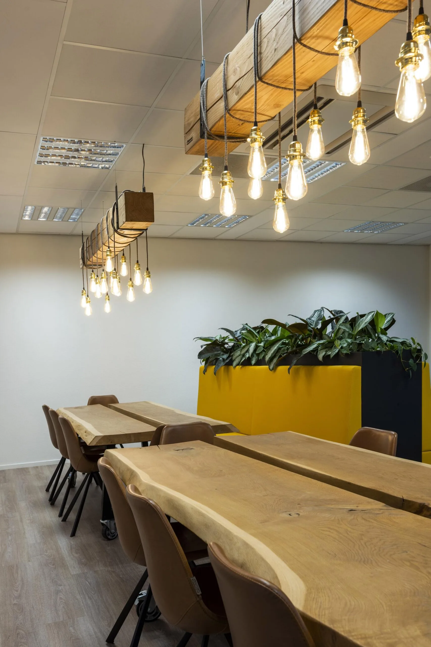 Moderne vergaderruimte met houten tafels, industriële hanglampen en een grote plantenbak in gele omkasting.