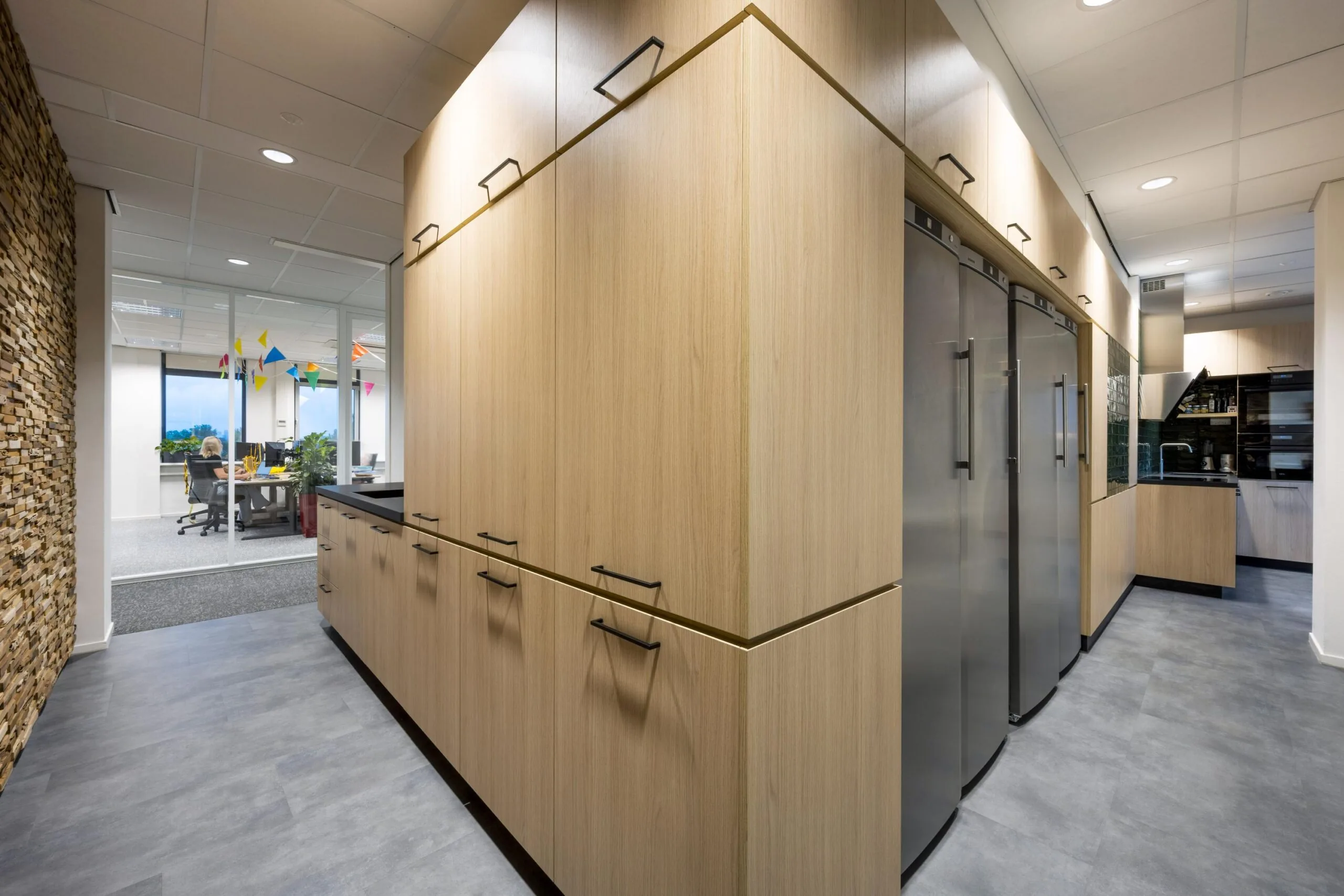 Moderne bedrijfskeuken met houten kasten, roestvrijstalen koelkasten en zicht op een kantoorruimte met kleurige slingers.