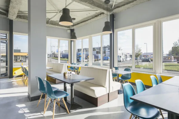 Modern en licht ingericht horecagedeelte aan de Hoefsmidstraat met blauwe stoelen, witte zitbanken en grote ramen met uitzicht op een industrieterrein.