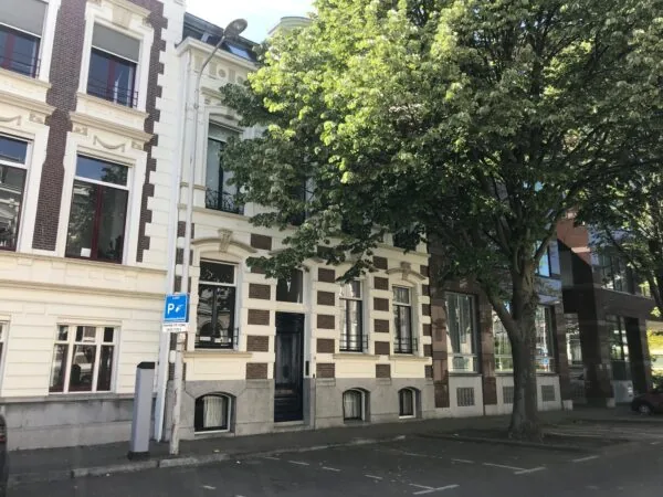 Voorgevel van een klassiek woongebouw aan de Burgemeester van Karnebeeklaan met bomen en parkeervakken op de voorgrond.