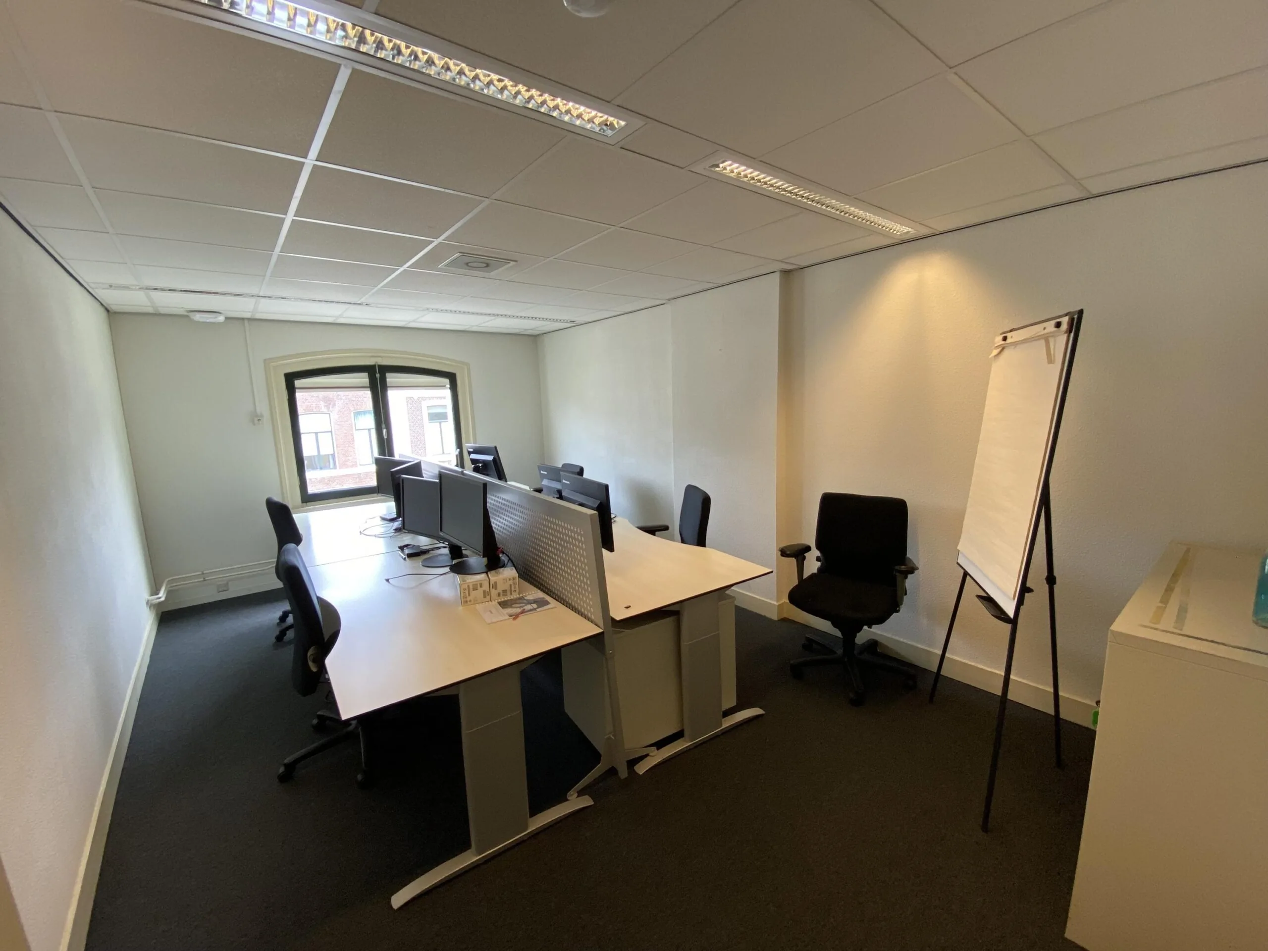 Kleine kantoorruimte aan de Burgemeester van Karnebeeklaan met meerdere bureaus, bureaustoelen, computerschermen en een flip-over.