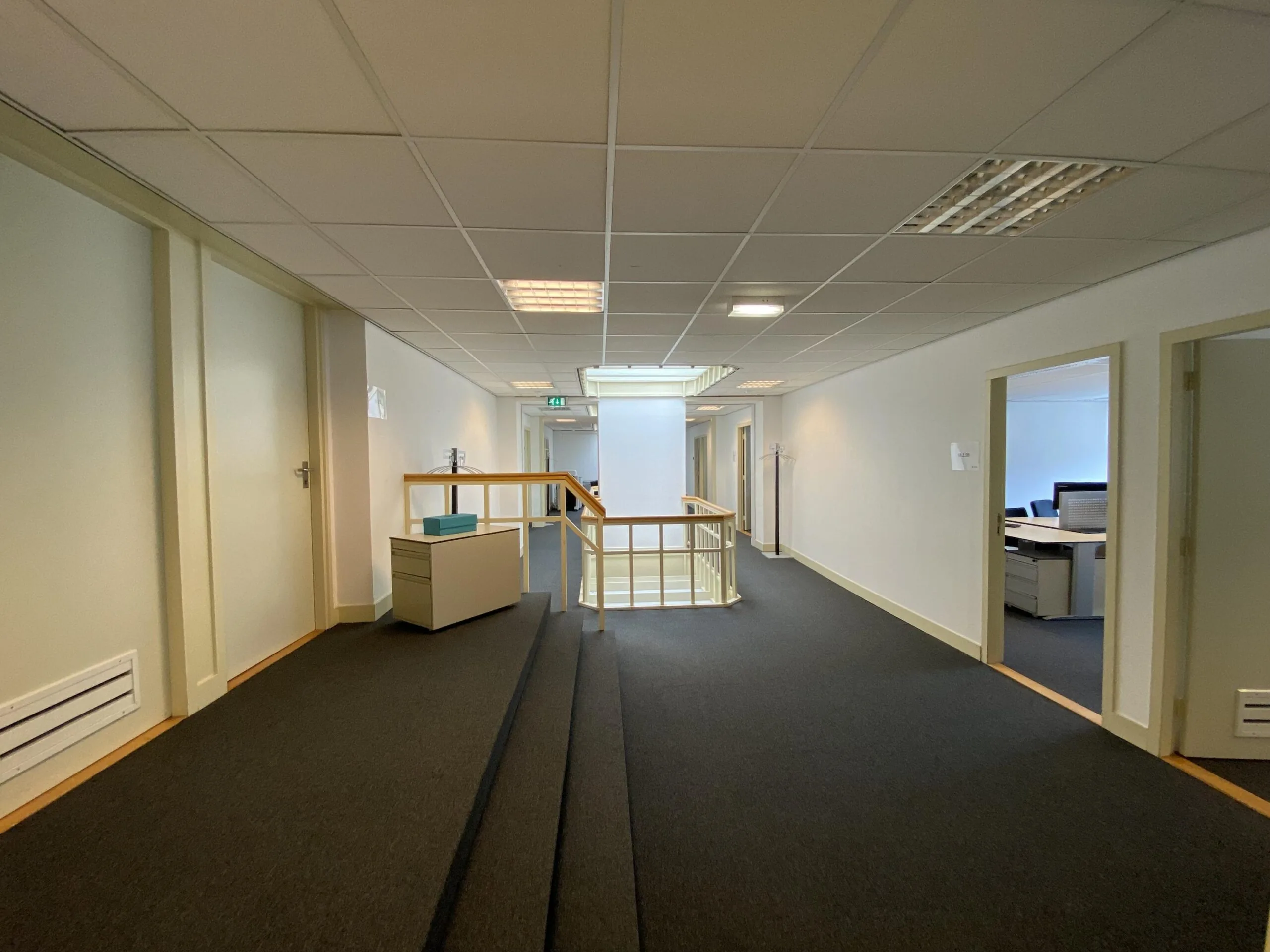Gang van een kantoorpand aan de Burgemeester van Karnebeeklaan, met open deuren naar kantoorruimtes en een trap met houten leuning.