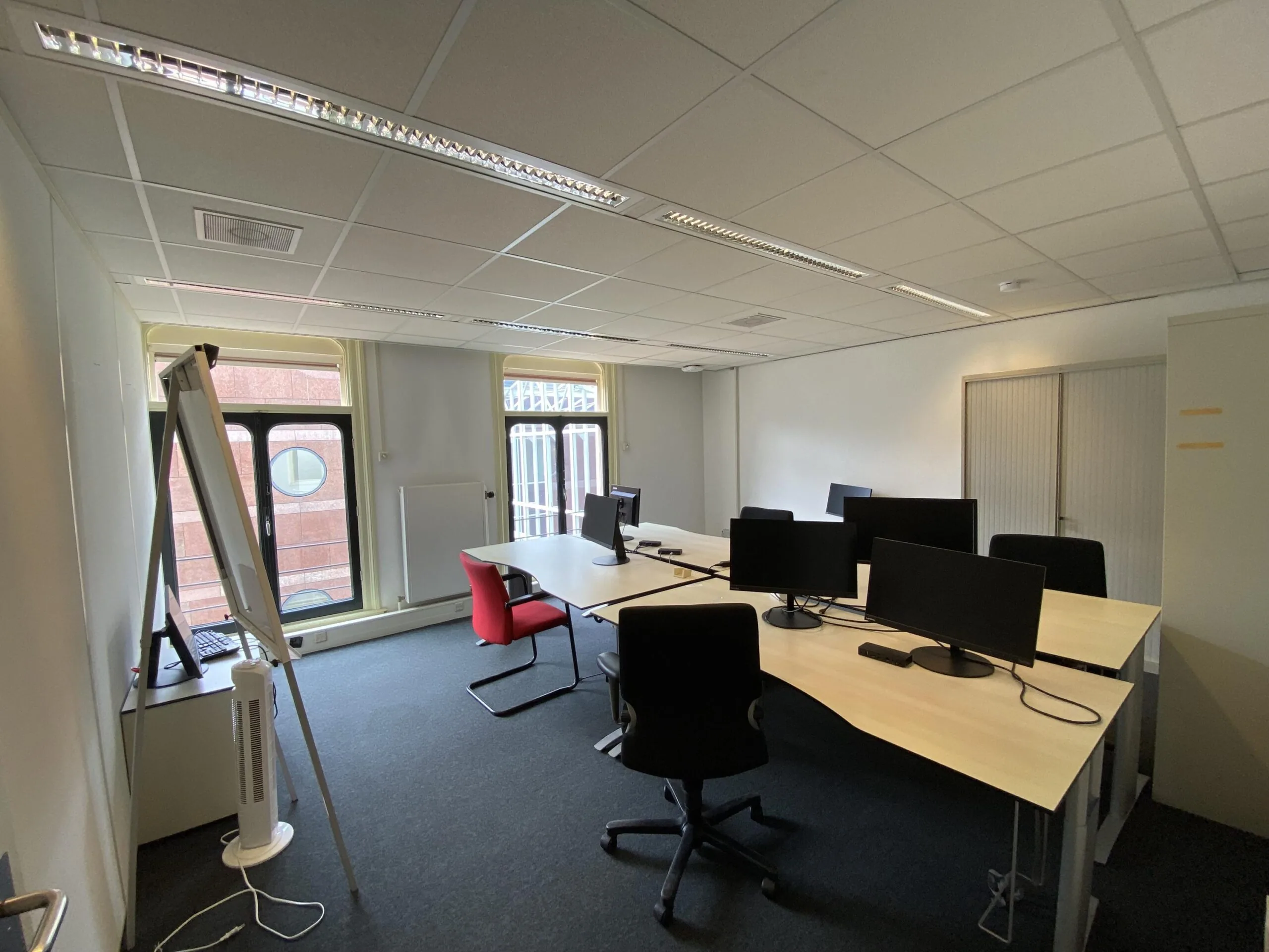Kantoorruimte aan de Burgemeester van Karnebeeklaan met bureaus, computers, stoelen en een flip-over bij het raam.