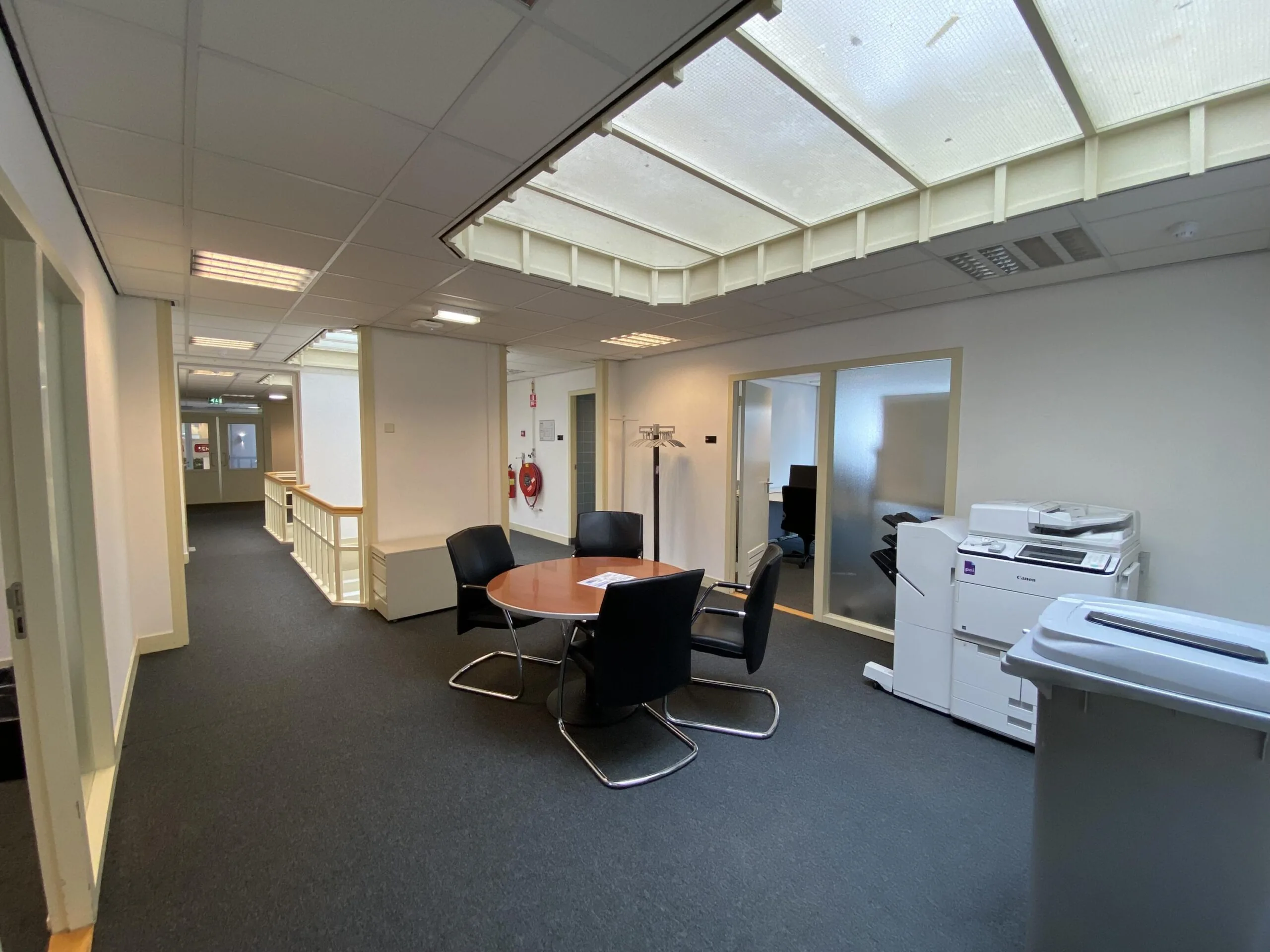 Kantoorruimte aan de Burgemeester van Karnebeeklaan met een vergadertafel, stoelen en een printerhoek onder een groot plafondraam.