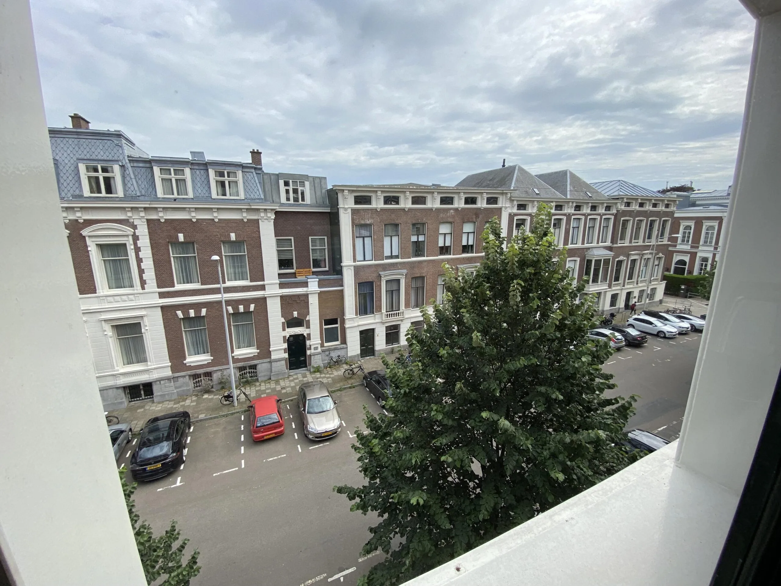 Uitzicht vanaf een hoger gelegen raam op de burgemeester Van Karnebeeklaan met geparkeerde auto's en historische herenhuizen.