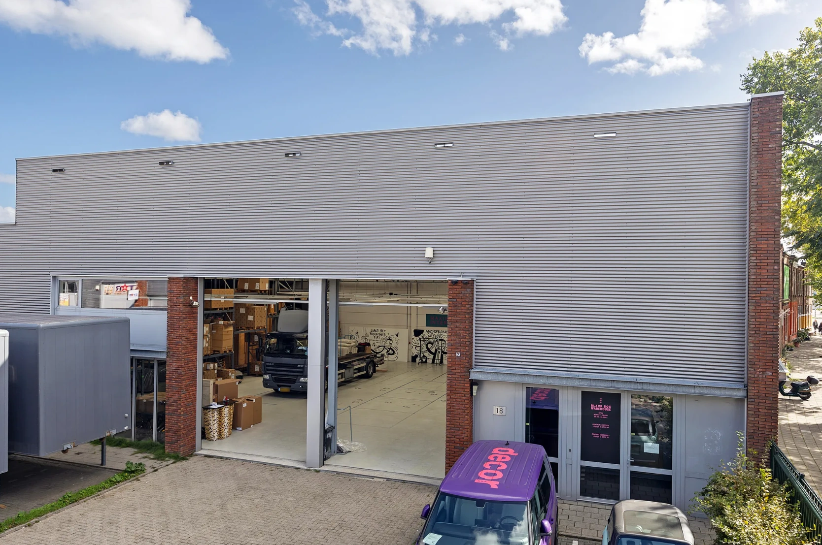 Bedrijfsgebouw aan de Schaafstraat met open loodsdeur, geparkeerde voertuigen en zicht op een magazijnruimte binnenin.