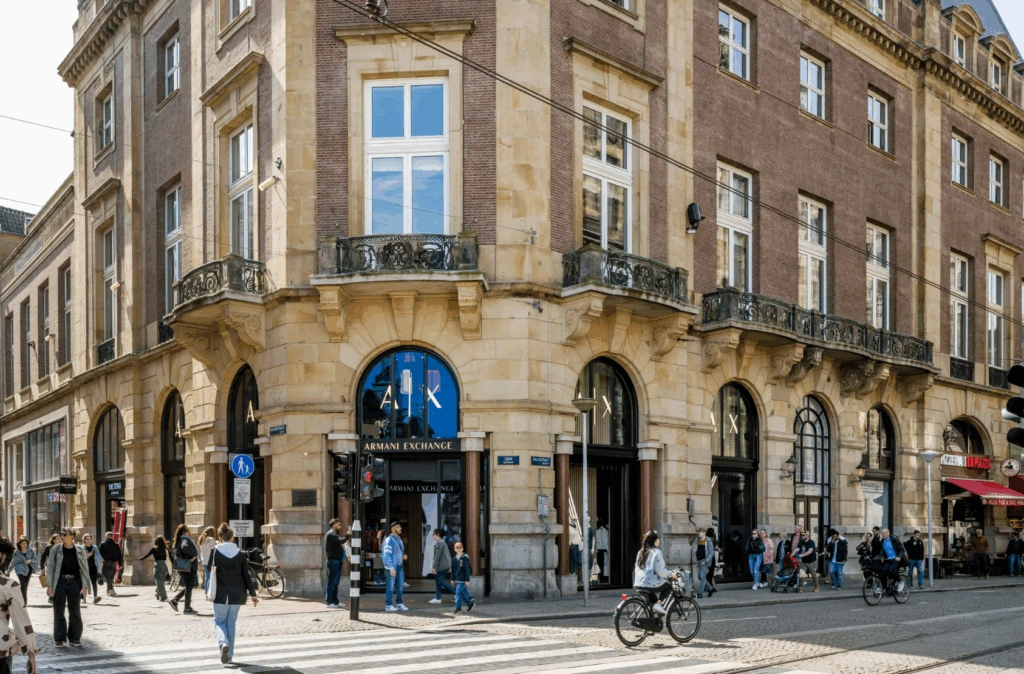 Drukke straat op de hoek van de Paleisstraat met winkels zoals Armani Exchange in een historisch gebouw.
