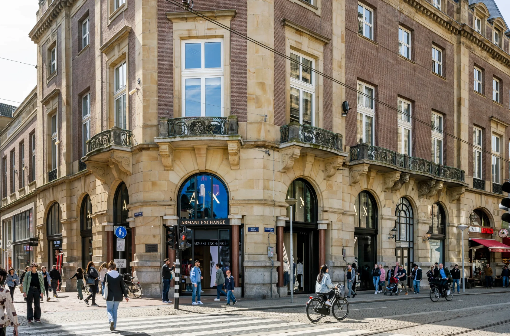 Drukke straat op de hoek van de Paleisstraat met winkels zoals Armani Exchange in een historisch gebouw.