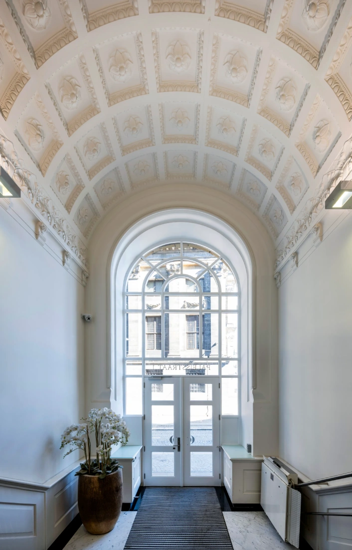 Ingang van een monumentaal pand aan de Paleisstraat met een hoge gewelfde plafonddecoratie en glazen dubbele deuren.