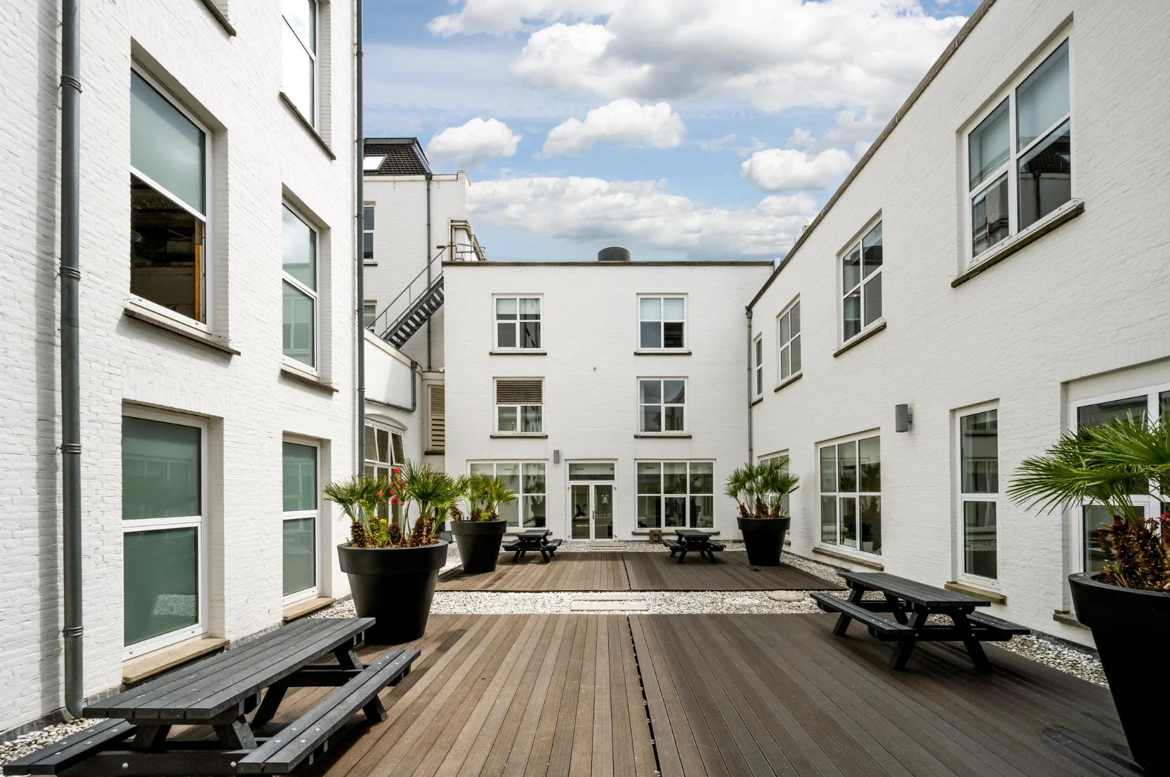 Binnenplaats van een modern wit appartementencomplex aan de Paleisstraat, met houten vlonders, zwarte picknicktafels en grote bloempotten met palmen.