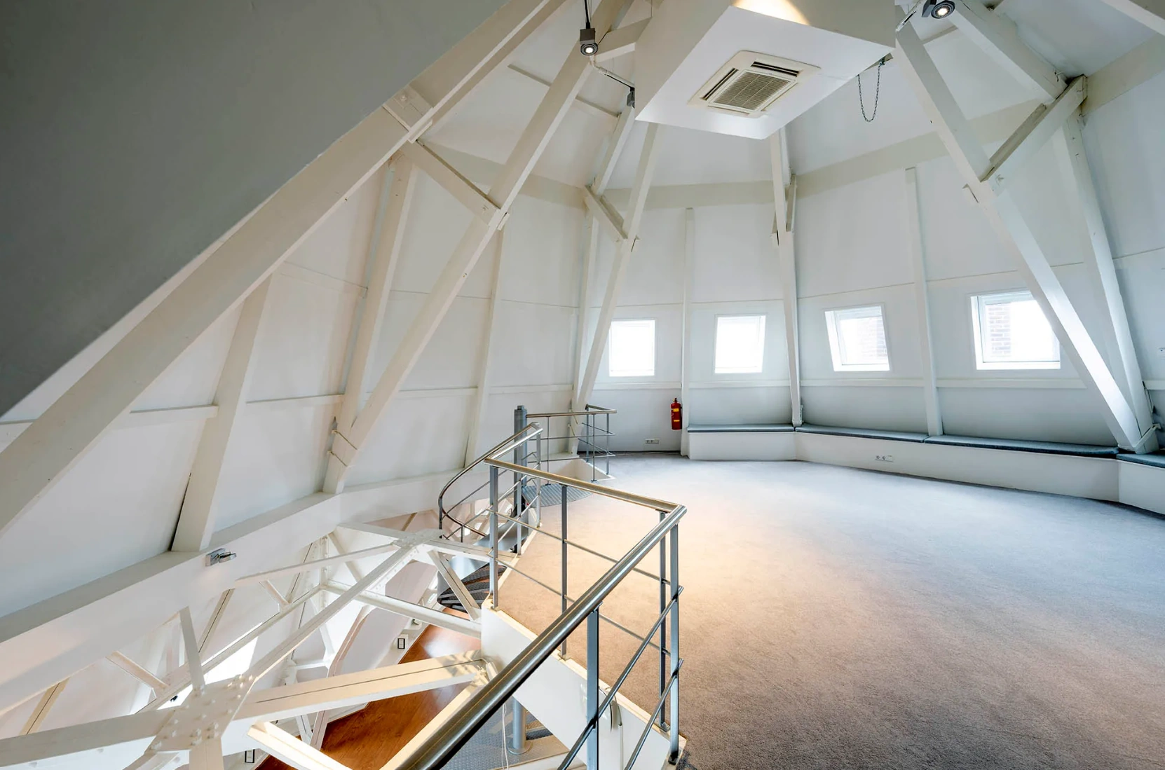 Ruime, lichtgekleurde zolderruimte met schuine wanden, ramen en een metalen trap in Paleisstraat.
