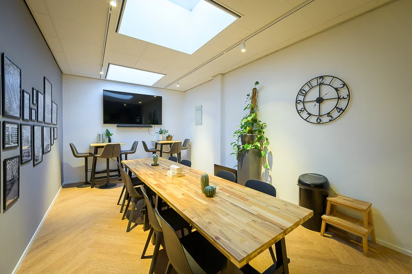Moderne vergaderruimte aan de Linschotenstraat met houten tafel, planten, grote klok en dakramen voor natuurlijk licht.