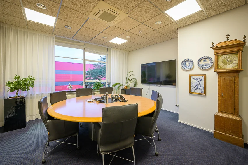 Vergaderruimte aan de Linschotenstraat met een ovale houten tafel, zwarte stoelen, een klok en wanddecoratie.