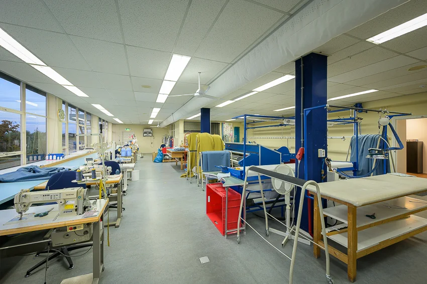 Atelier aan de Linschotenstraat met naaimachines, strijkapparatuur en werktafels in een ruime, goed verlichte werkruimte.