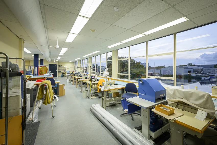 Naaiatelier aan de Linschotenstraat met werkplekken, naaimachines en grote ramen met uitzicht op buiten.