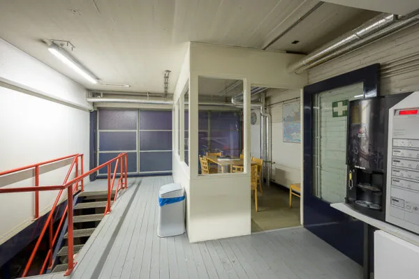 Kleine bedrijfs- of kantoorpauzeruimte met tafels, stoelen, koffieautomaat en een trap met rood leuningwerk.