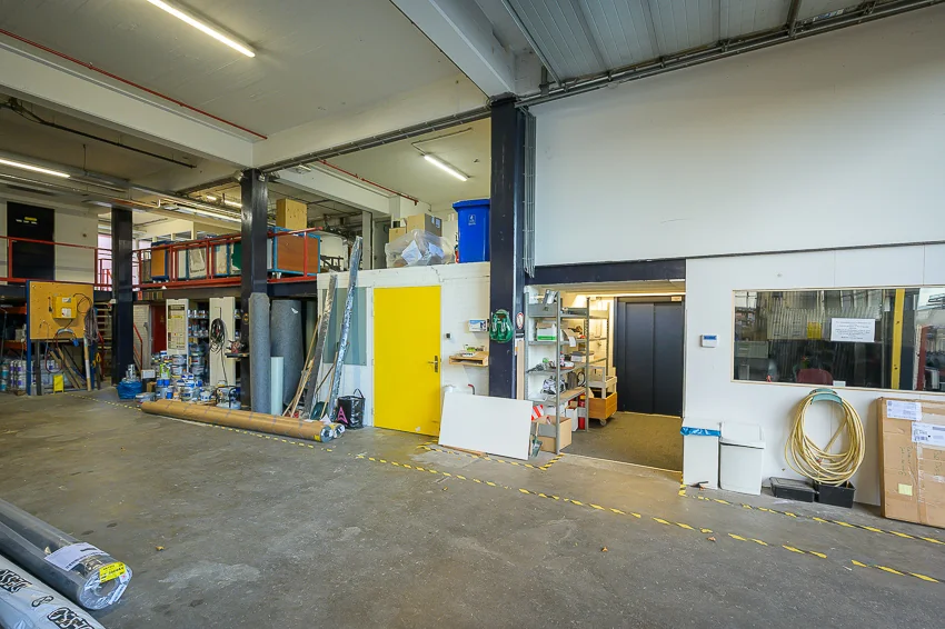 Binnenzicht van een industriële werkruimte aan de Linschotenstraat met opslagrekken, een gele deur en een kantoorruimte.