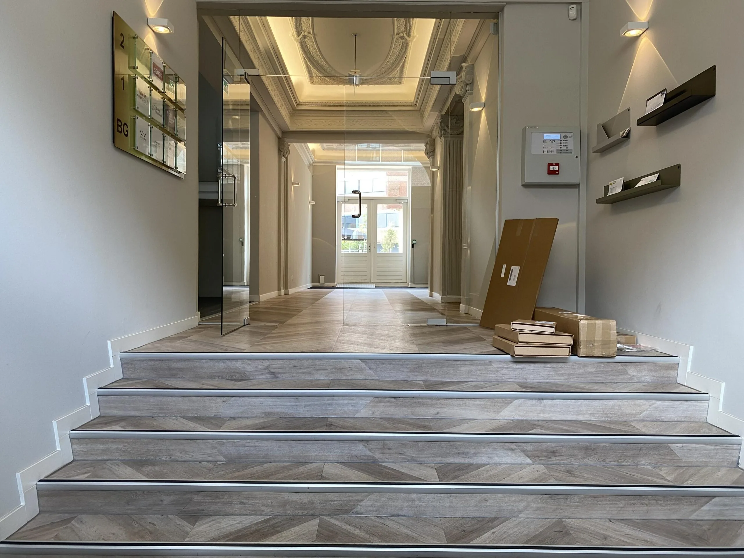 Ingangshal van een kantoorpand aan de Burgemeester van Karnebeeklaan met traptreden, een glazen deur en enkele bezorgde pakketten.