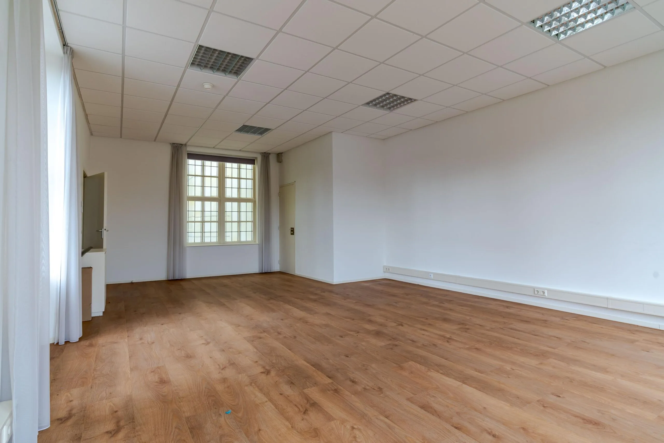 Leeg kantoor- of praktijkruimte aan het Lange Voorhout met houten vloer, systeemplafond en grote raampartij.