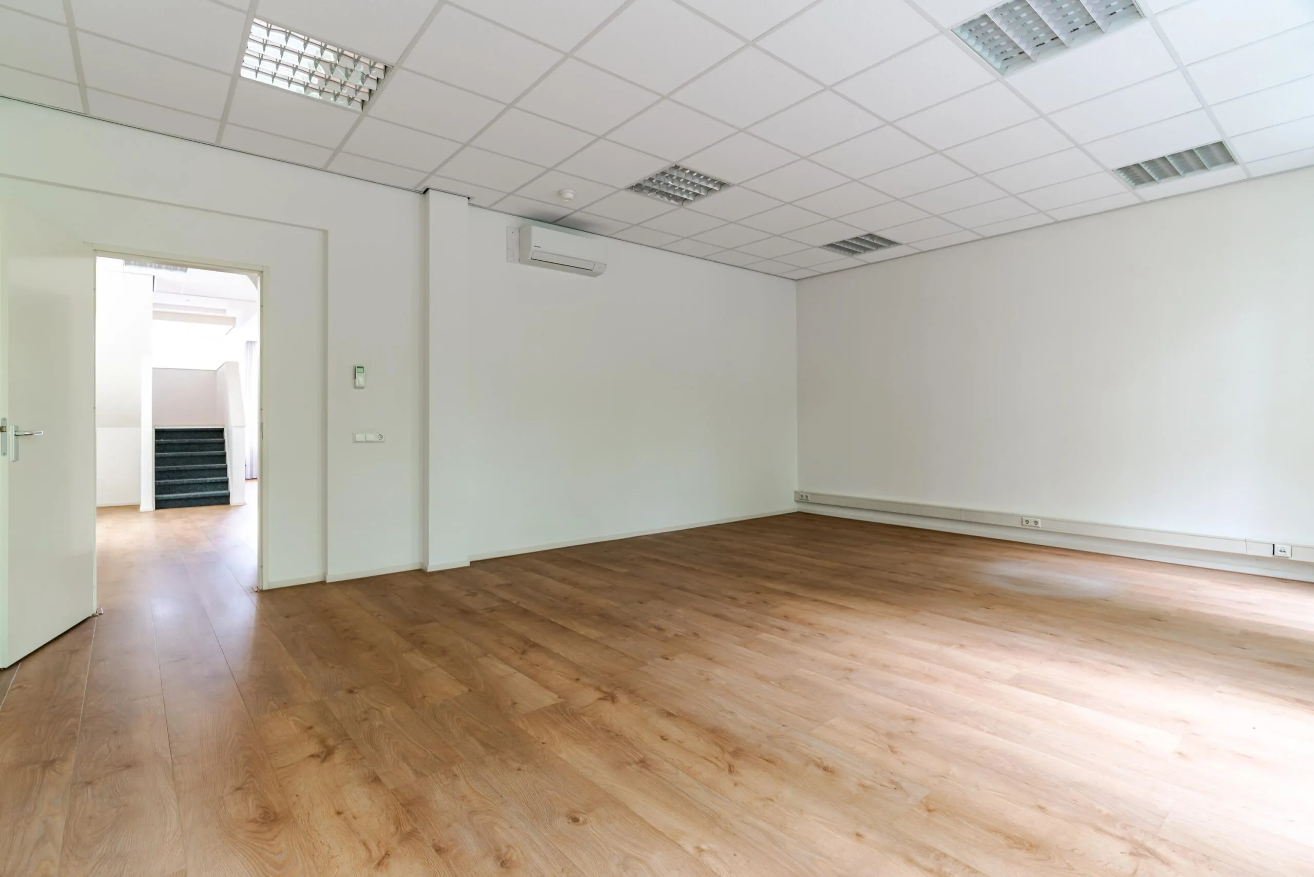 Leeg kantoorpand met houten vloer, systeemplafond en open deur naar een trapopgang.